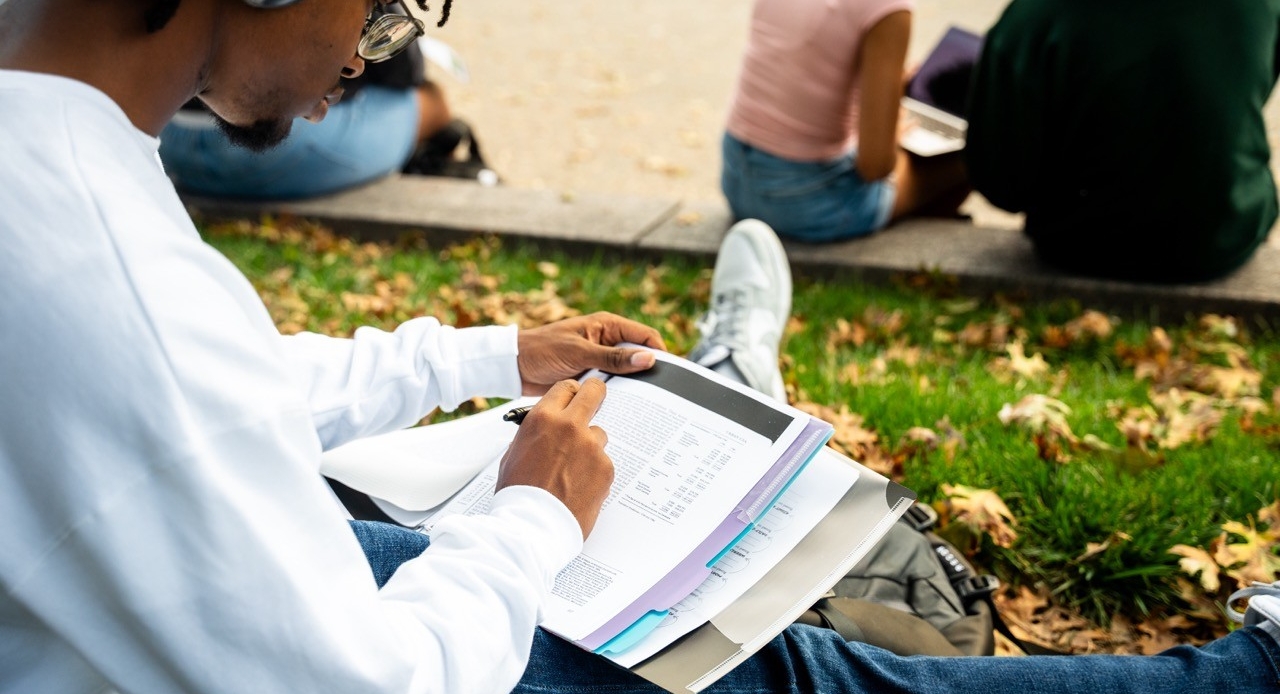 Student pictured from the back writing in a textbook outside on campus