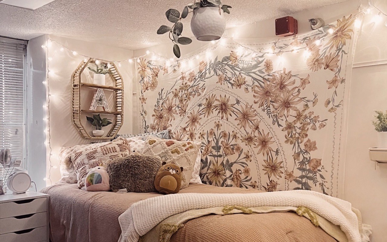 Bed and linens in tan and brown, with flower tapestry on wall, wicker shelf on wall, and pot with plant hanging from ceiling.