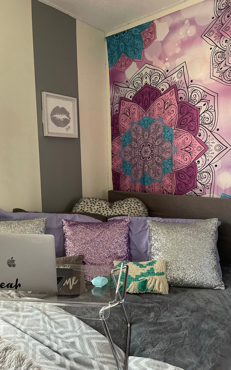 Apartment bed decorated in shades of gray and purple with pillows and a decorative wall hanging
