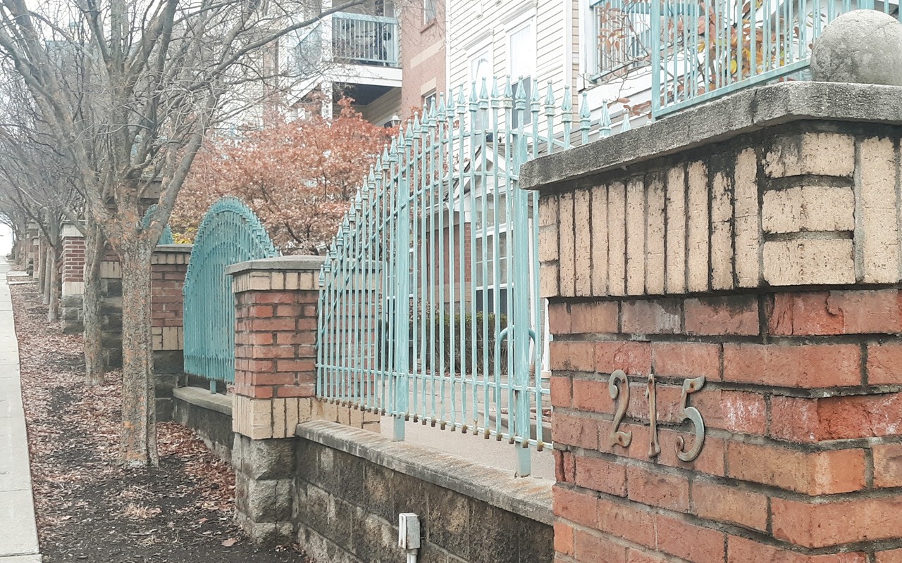 Front gate made of green iron railings with brick blocks in between at Bellevue Gardens. There is a line of trees next to the sidewalk.