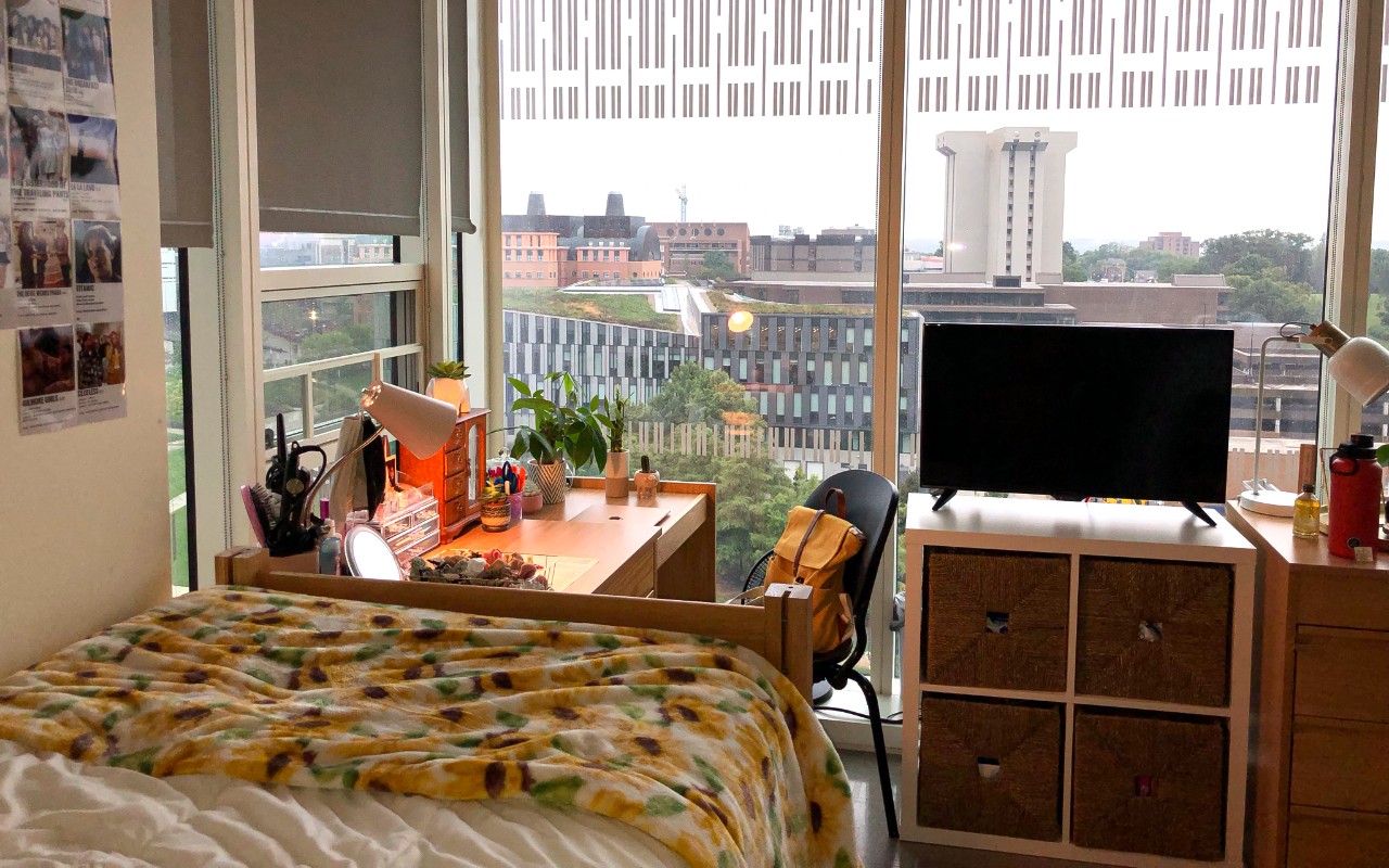 Large windows open the room to a view of campus. The edge of a twin bed, a television, and a desk are located in the room.