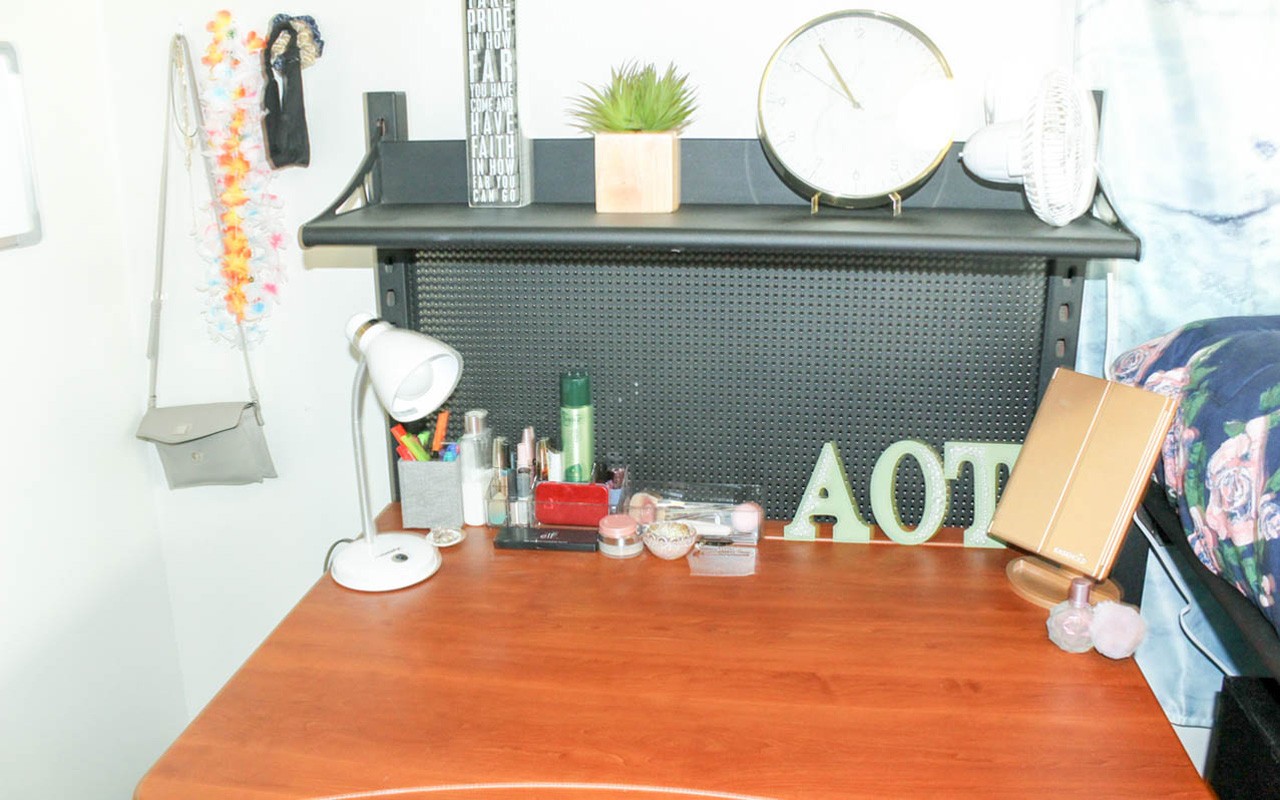 Desk with a shelf in a double room in a Stratford Heights suite. There is a bed to the right of the desk and a purse hanging on the wall to the left of the desk. There is a lamp and other personal items on the desk. There is a clock and other items on the shelf above the desk.
