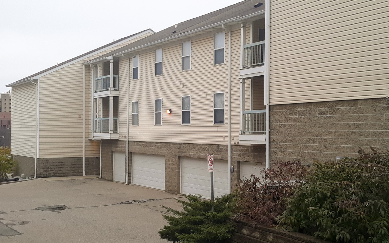 Exterior of Bellevue Gardens apartments. with a yellow fire hydrant  embedded in the public sidewalk. The ground floor apartments have a concrete patio. There is a green iron and brick gate that meets the sidewalk in front of the property And a row of trees in front of the gate..