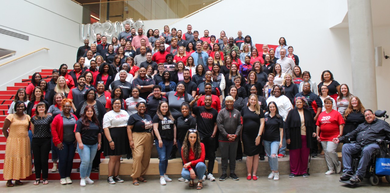 Over 100 staff members dressed in red and black stand on stairs in the Lindner College of Business Atrium