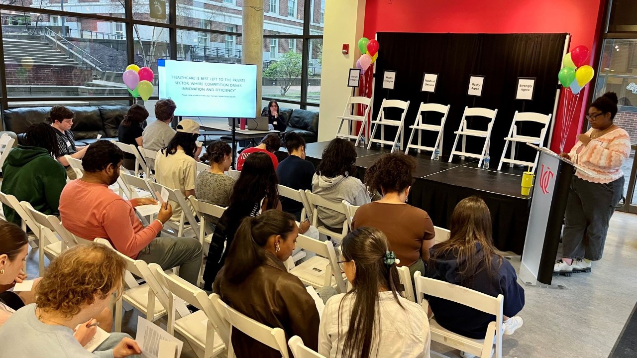 Students are attending one of UC Votes' Dinner & Dialogue Events. There are students sitting in the audience, on a stage, and one student speaking from a podium. 