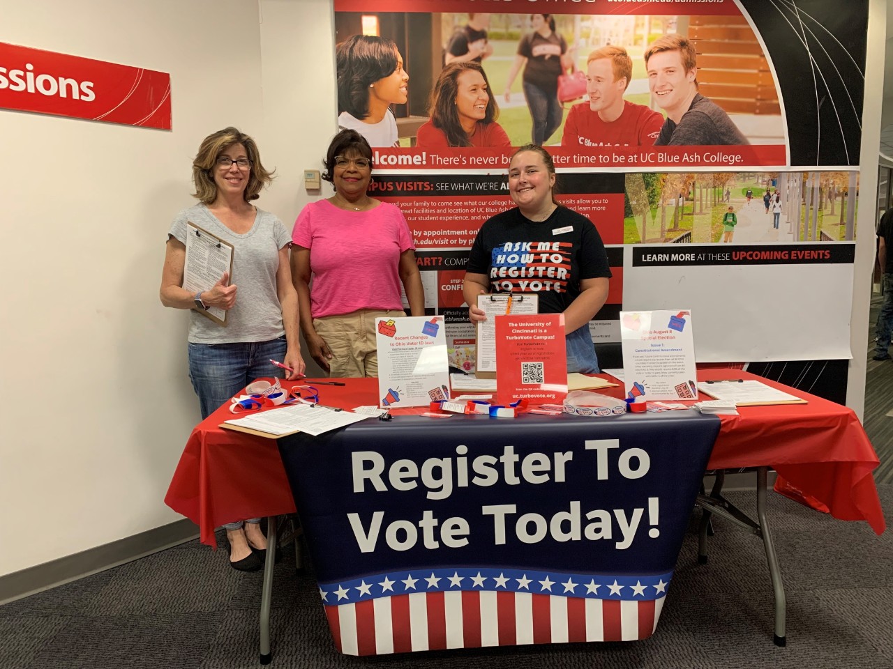 A UC Votes voter registration table at UC Blue Ash. There is one student associate working alongside two community volunteers. 