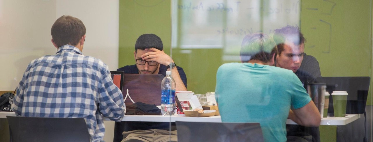 A student reads from a text book, holding a dry erase marker with his back to a math problem on the board. 