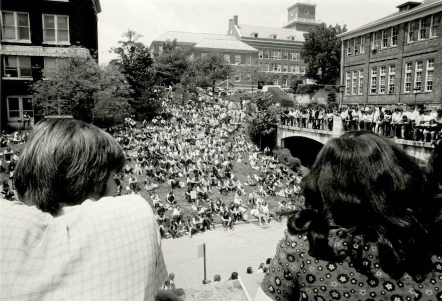 Black and white photo of the backs of two heads looking onto a lawn and bridge filled with students