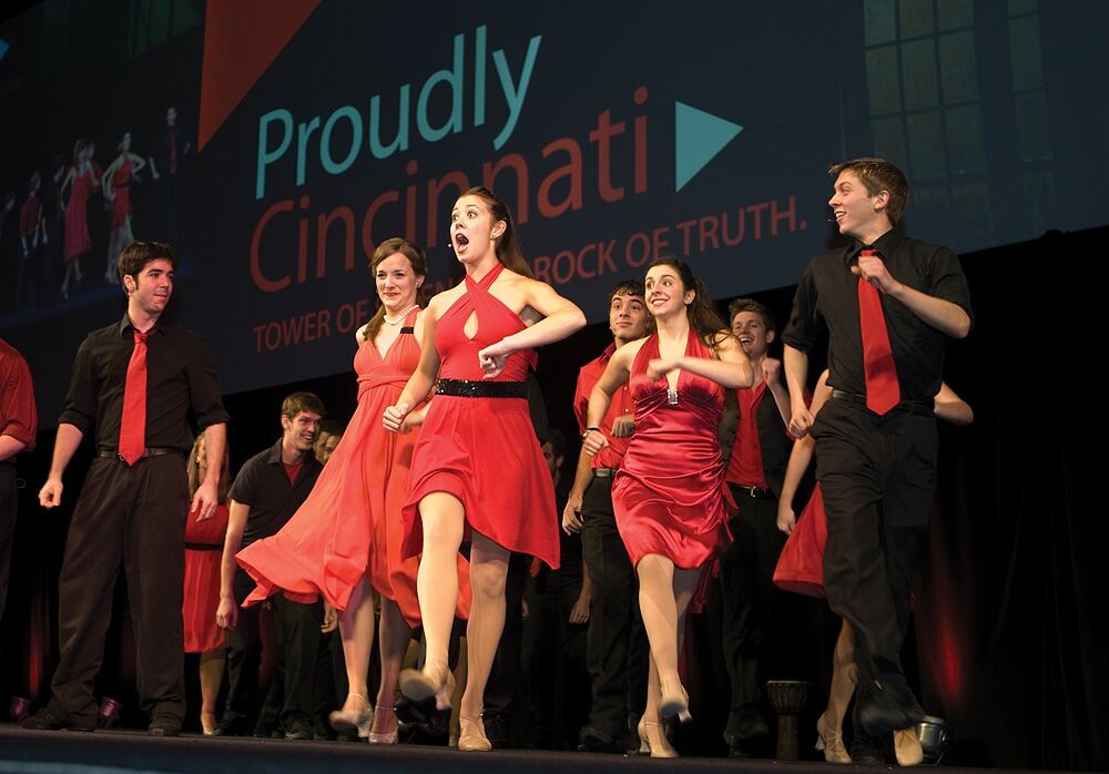 Students sing and dance onstage in front of a Proudly Cincinnati sign