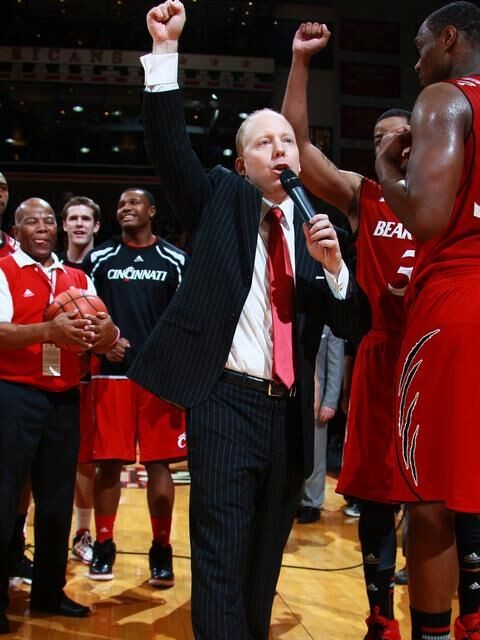 Coach Mick Cronin speaks on a microphone with hand raised around basketball players on court