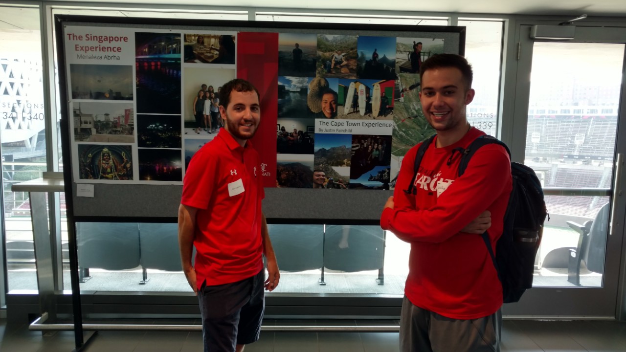 Two UC students wearing red UC shirts pose in front of a poster with several images from a past trip to Cape Town, South Africa.