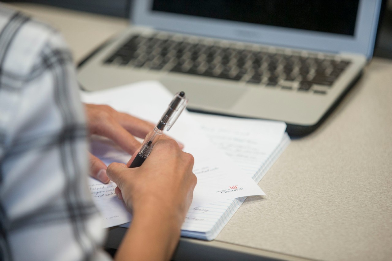 A closeup of a person at a desk writing in a notepad with a laptop computer in the background