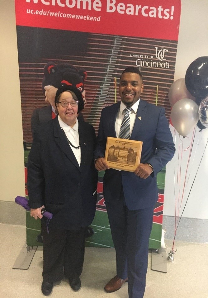 Smiling male student in graduation cap, gown, and many honors tassels stands outside Nippert Stadium, holding a diploma, with his other arm around a woman