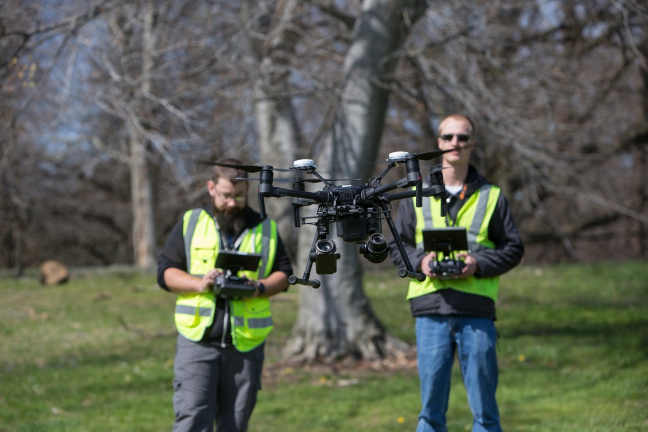 two young men stand outside wearing neon yellow safety vests, using remote controls to pilot a drone in the foreground