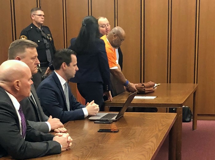 Men in suits sit in a court room table while a defendant in an orange jump suit stands surrounded by his attorney at another courtroom table in a courtroom
