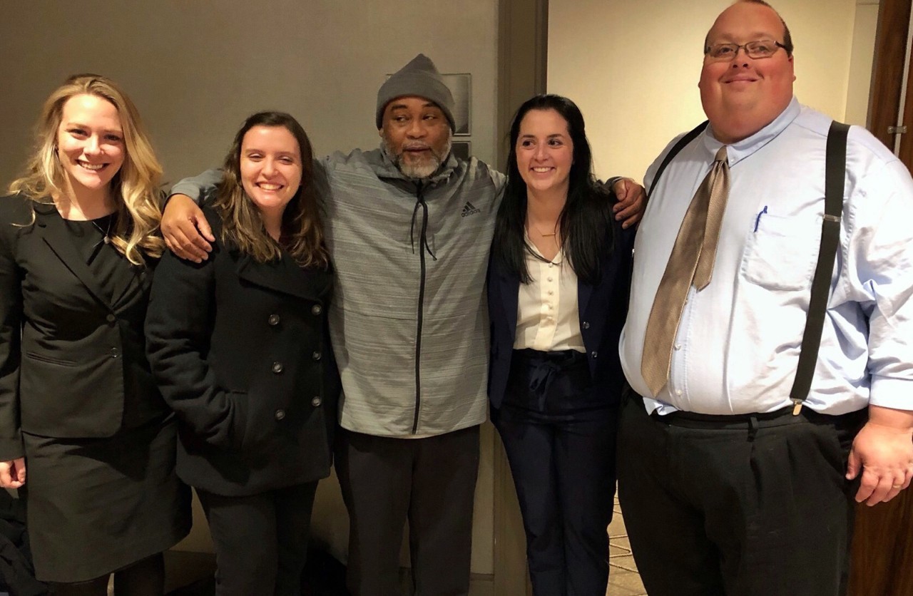 Five people stand together with their arms linked around each other's shoulders smiling at the camera shoulders