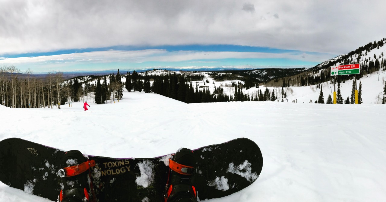 A view of a snowy, tree-lined ski slope on a bright day, taken from a point of view that shows the photographer's feet in a snowboard. The skies are blue with clouds and there is only one other person on the slope