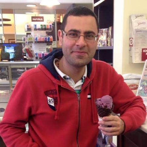 A young adult man with short dark hair and wire frame rectangular glasses, wearing  a red hooded sweatshirt, stands in an ice cream parlor holding an ice cream cone with purple ice cream on it