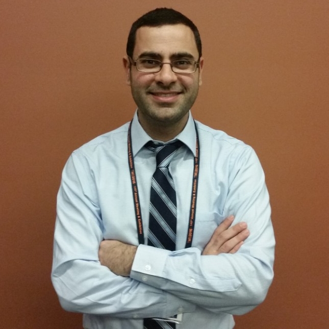 A young adult man with short dark hair and wire frame rectangular glasses, wearing a white button down shirt and blue striped tie, arms folded across his chest, against a brownish orange wall