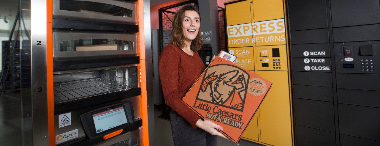woman holding a Little Caesars Pizza box in front of a pizza warmer with keypad for PIN entry