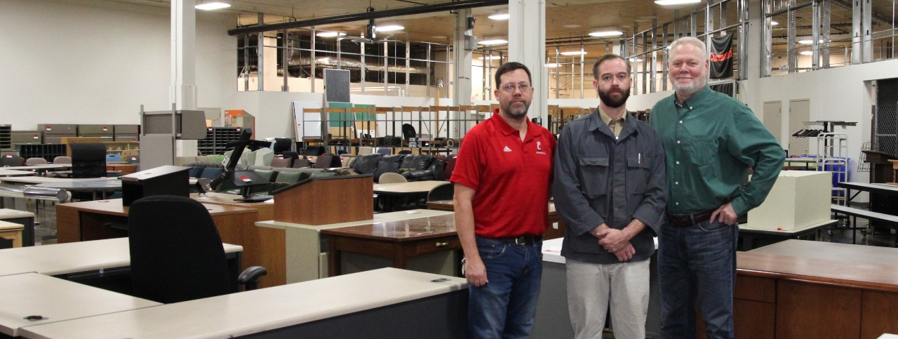 Three men stand among UC's Surplus merchandise in their Fishwick warehouse location.