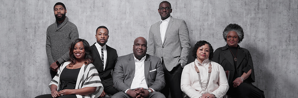 Seven adults in monochromatic business dress sit and stand in three rows in front of a grey wall