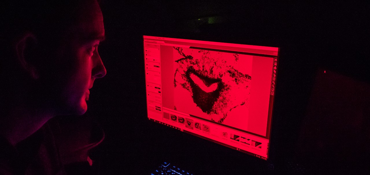 UC professor Nathan Morehouse looks at a computer monitor with a red filter that reflects red light on his face. 
