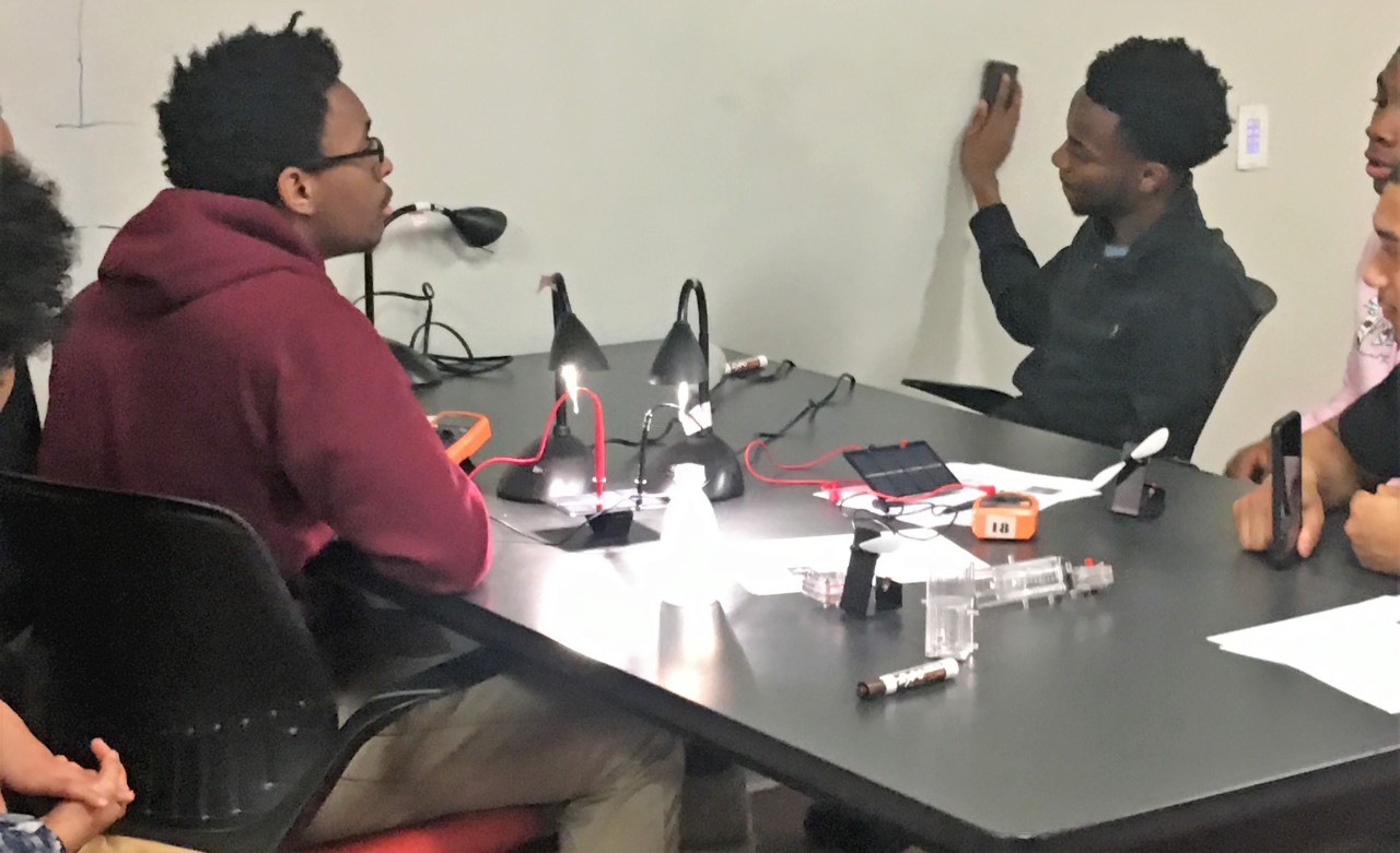four young people sit at a table, holding smart phones. There are lights, voltmeters, and electronic devices and wires across the table. They seem to be using the devices to text an electrical device