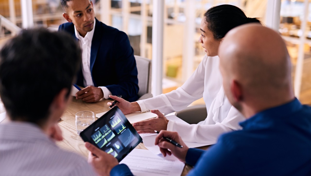 Several adults hold a discussion at a meeting room, one of whom is holding a tablet with an infographic on display