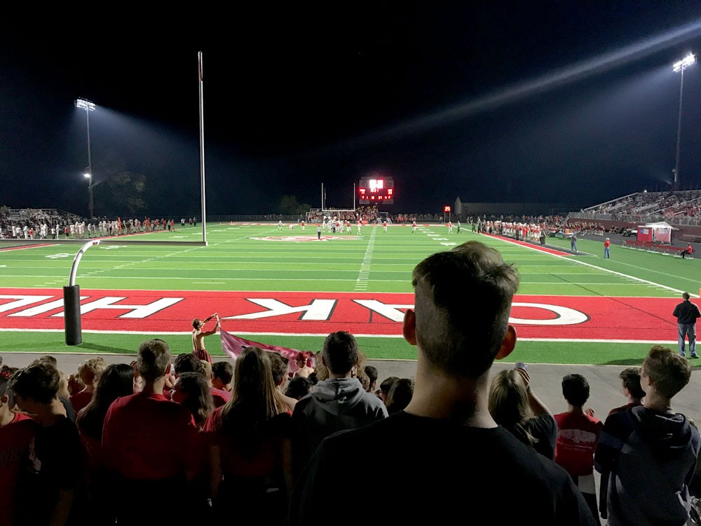 Teenaged boy stands in a crowd facing a football field at night