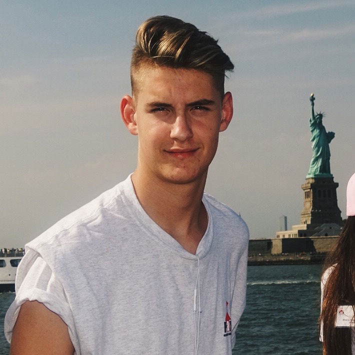 Teenager in light gray shirt smiles in front of the Statue of Liberty
