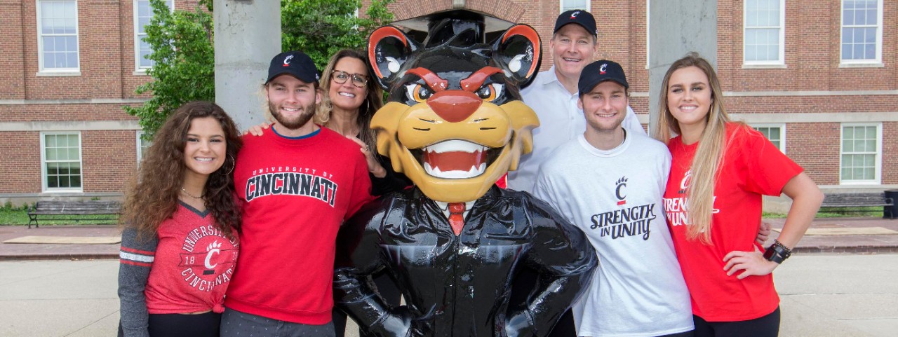 A family of six gathers around a statue of the UC Bearcat dressed in graduation clothing outside of McMicken Hall. 