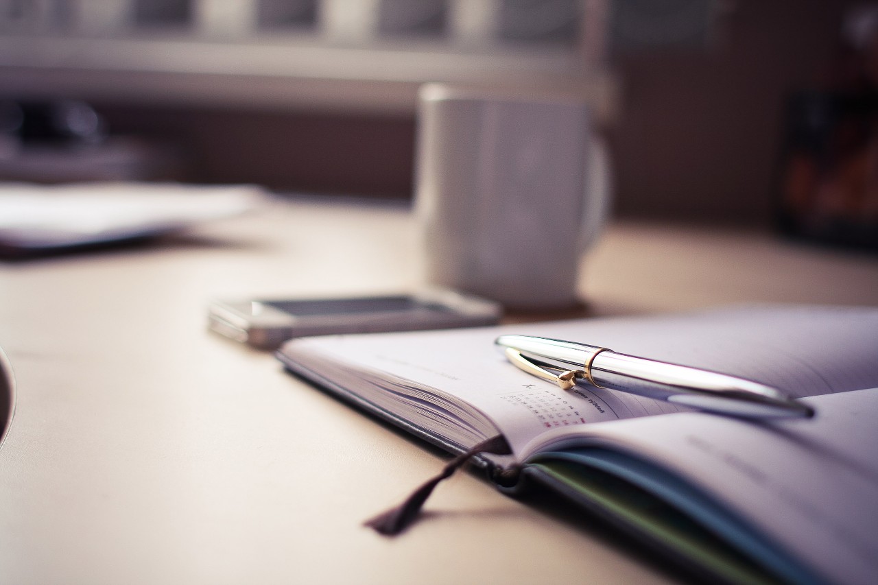 Ink pen laying on top of calendar with phone and mug on background on desk.