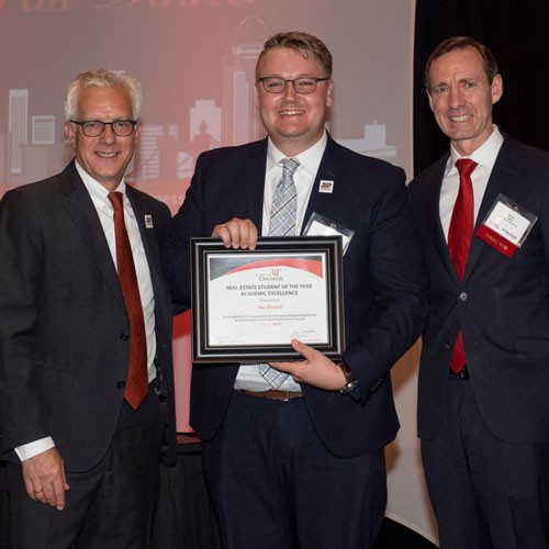Three men stand and smile and the middle one holds a framed award