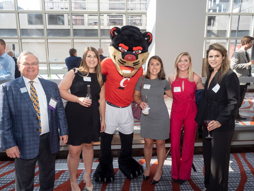 Dinner guests in business professional attire stand and smile next to bearcat mascot