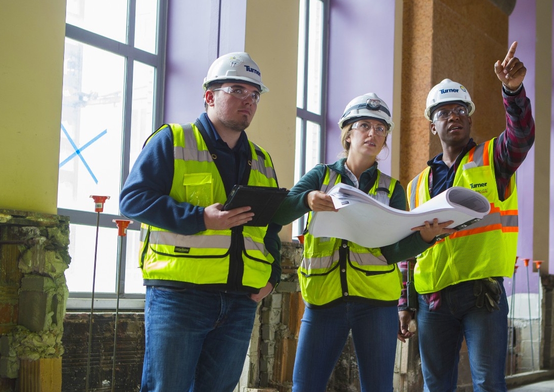 Three people in safety vests and hard hats hold blueprints while pointing at a job site.
