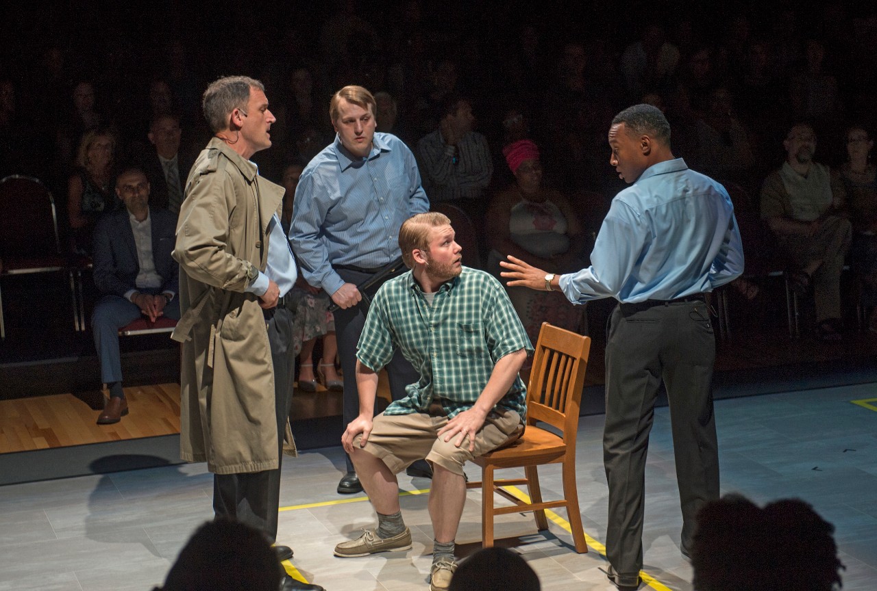 Stage scene of man in chair being interrogated by three other men