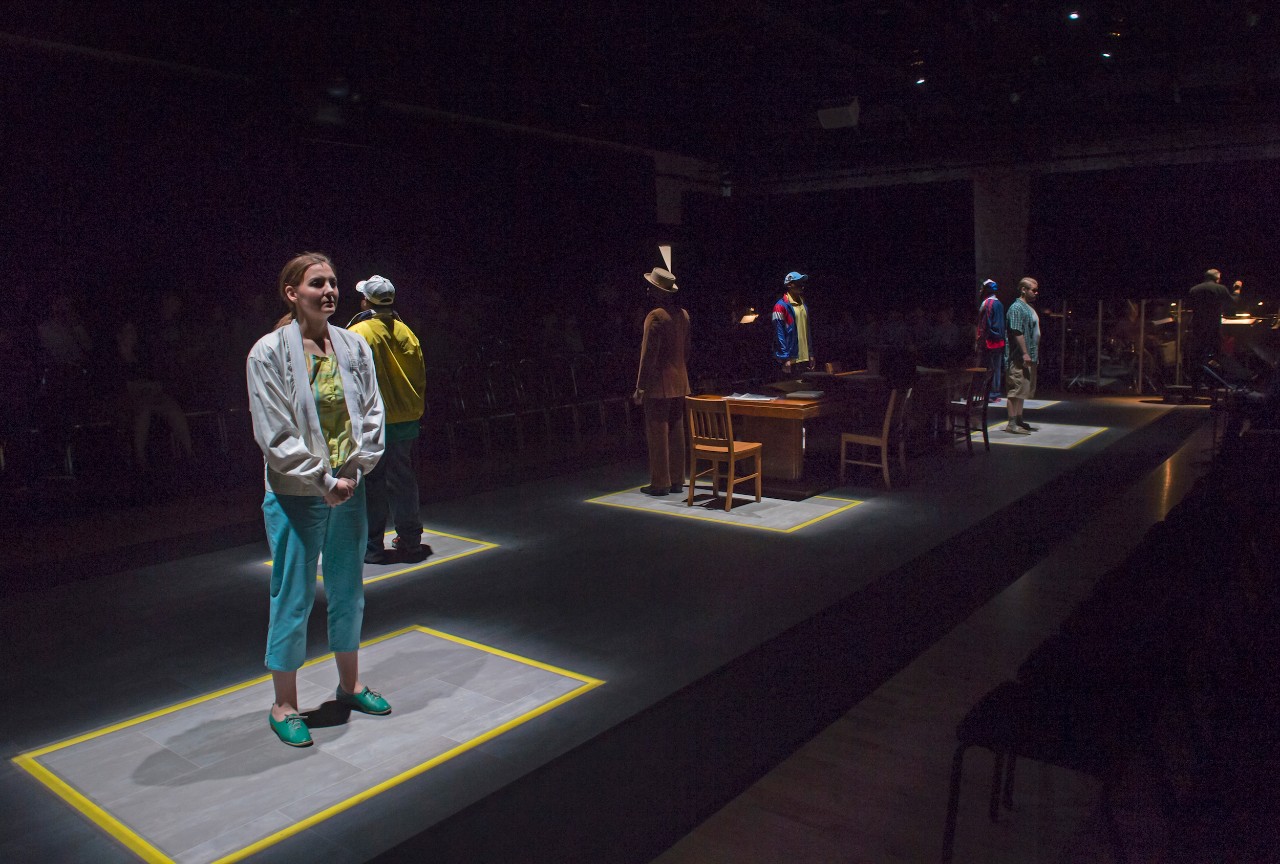 Six people on stage standing in squares lit with dramatic lighting