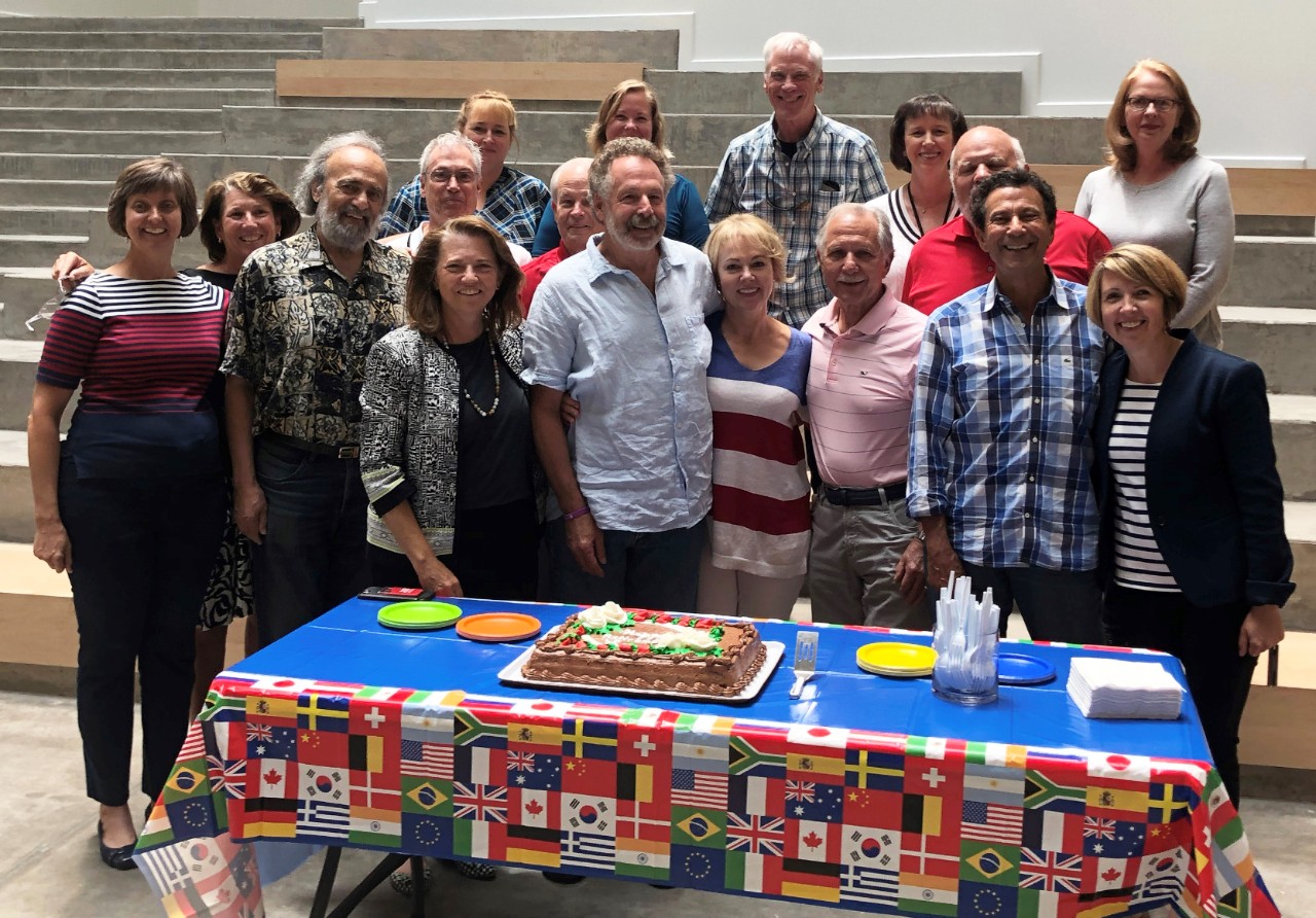 People pose with a retirement cake on a table decorated with international flags.