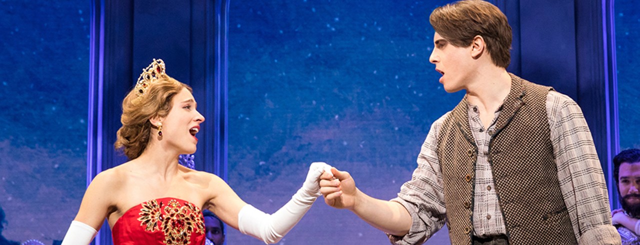 Woman in a gown, gloves and crown holds hand with a man in a theater production