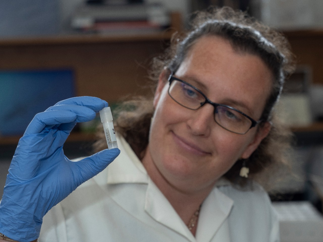 Brooke Crowley in a labcoat and gloves holds up a vial.