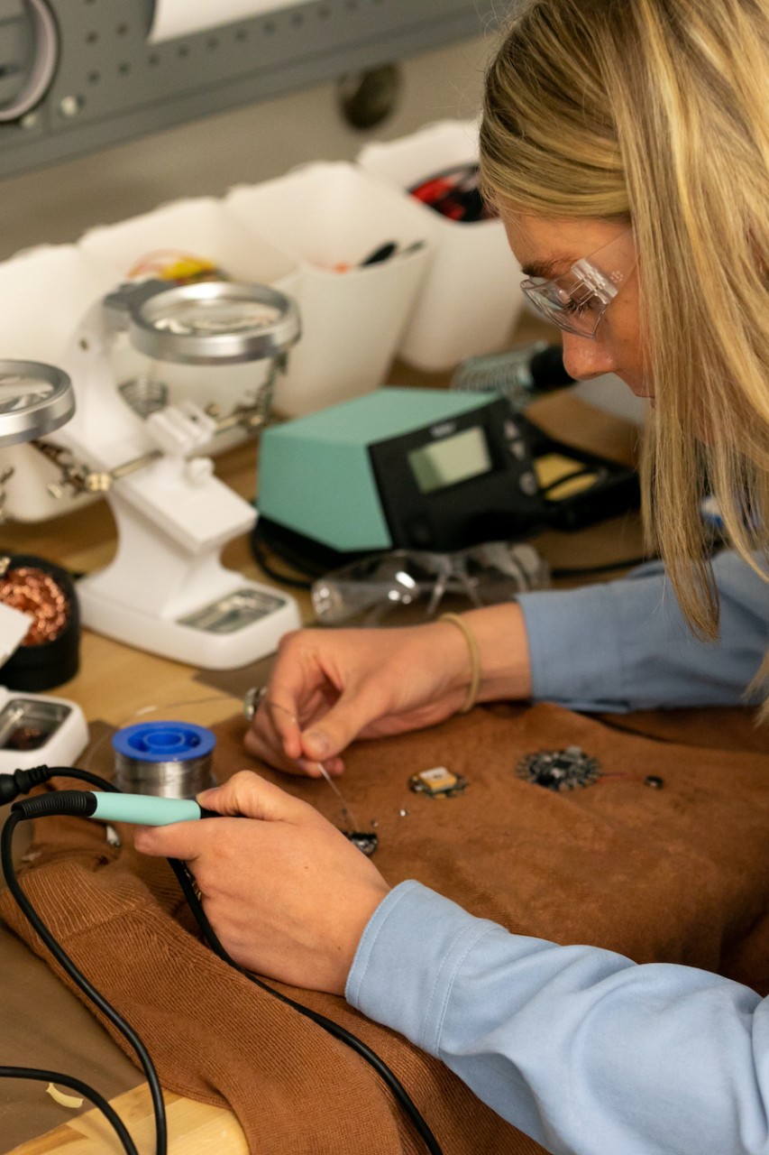 Woman at a work station uses soldering equipment to embed wearable tech into a brown sweater