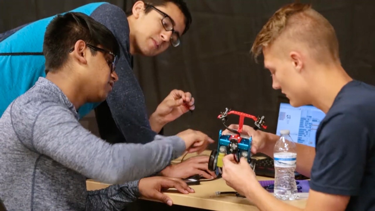 Three students work on a small handheld robot with wheels.