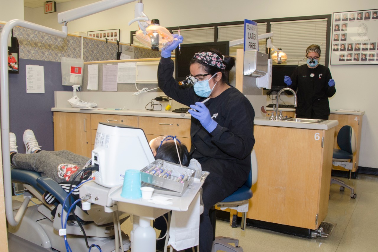 UC Blue Ash dental clinic. Patient in chair getting their teeth worked on by a hygienist. Another hygienist is standing  off to the back.