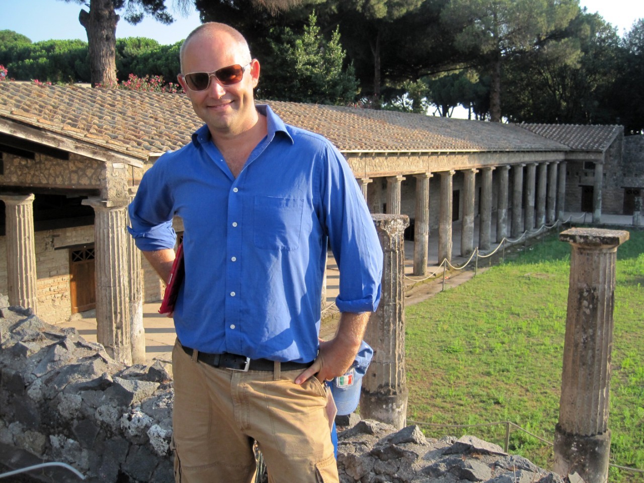 a man wearing a blue shirt and tan pants stands in front of roman ruins