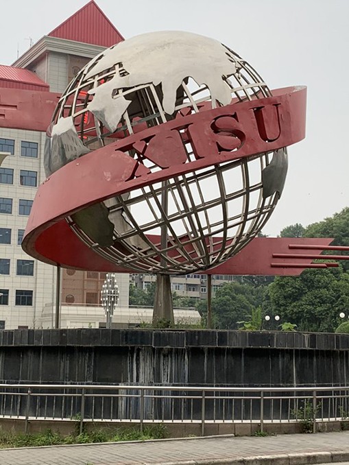 Outdoor art: metal sculpture of a globe with a banner reading XISU; building in the background