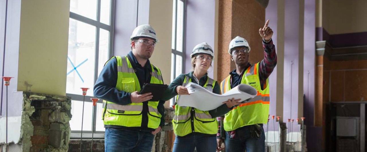 UC co-op students in hardhats and safety vests look over blueprints.