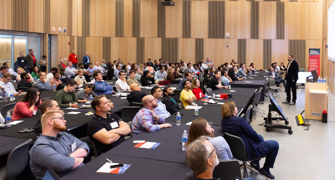 Symposium attendees sit in large conference room watching a man give a presentation