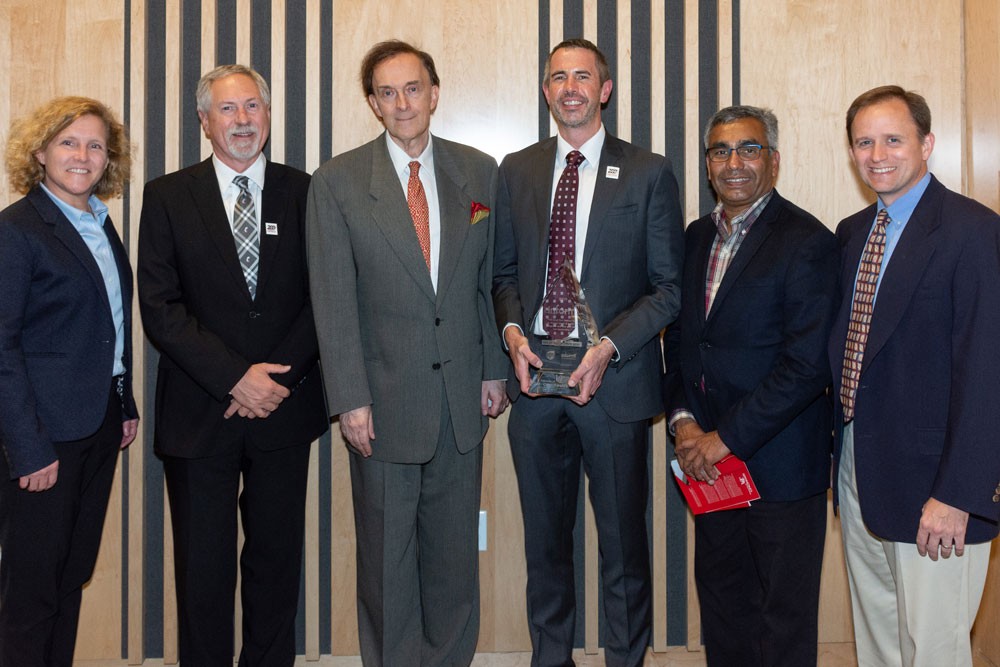 one woman and five men stand and smile wearing suits. One man holds a large glass award