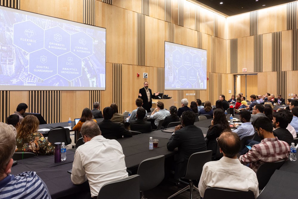 a man stands in front of two large screens giving a presentation in a crowded conference room
